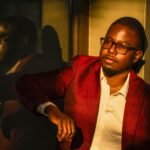 Stylish man in red jacket reflects in window light for dramatic portrait.